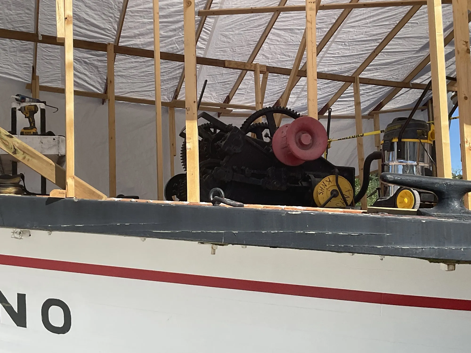 Black geared winch with a red drum on the forward main deck, behind a cleat, under the temporary shelter