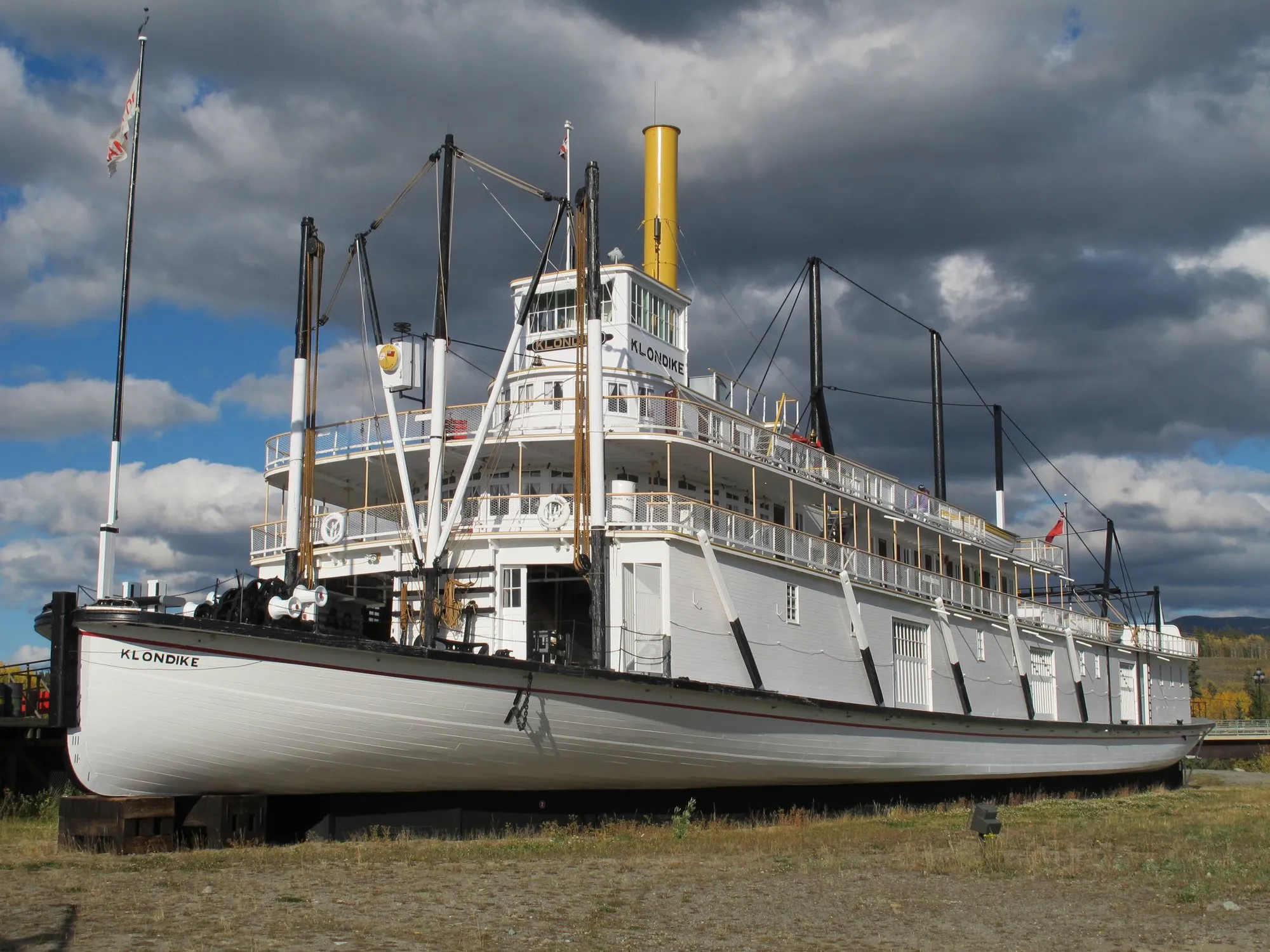 The Klondike on blocking beside the river at Whitehorse, white hull and decks, yellow stack, under dark clouds with autumn trees behind