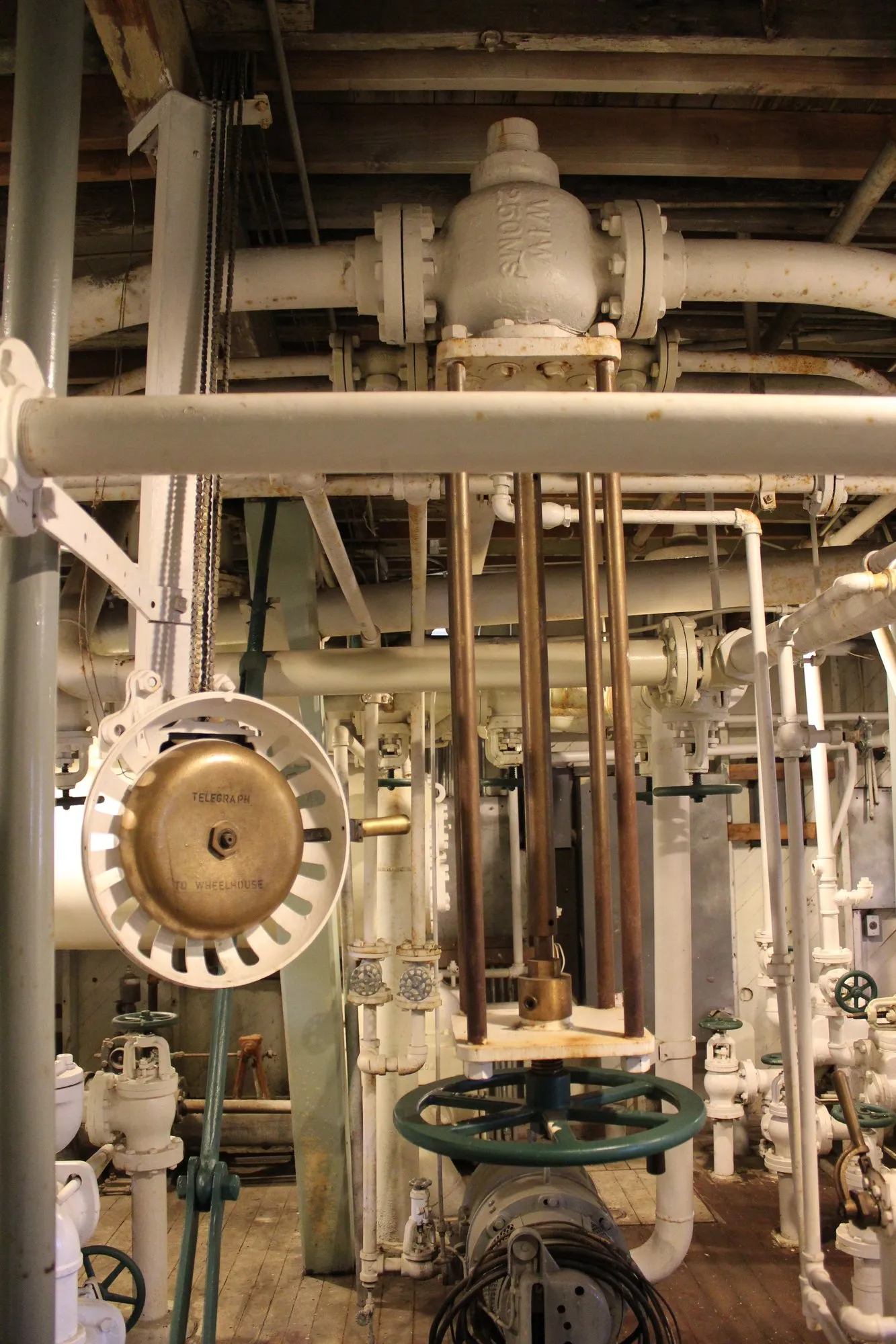 Engine room controls with a brass signal bell, vertical control rods and a valve handwheel among white piping