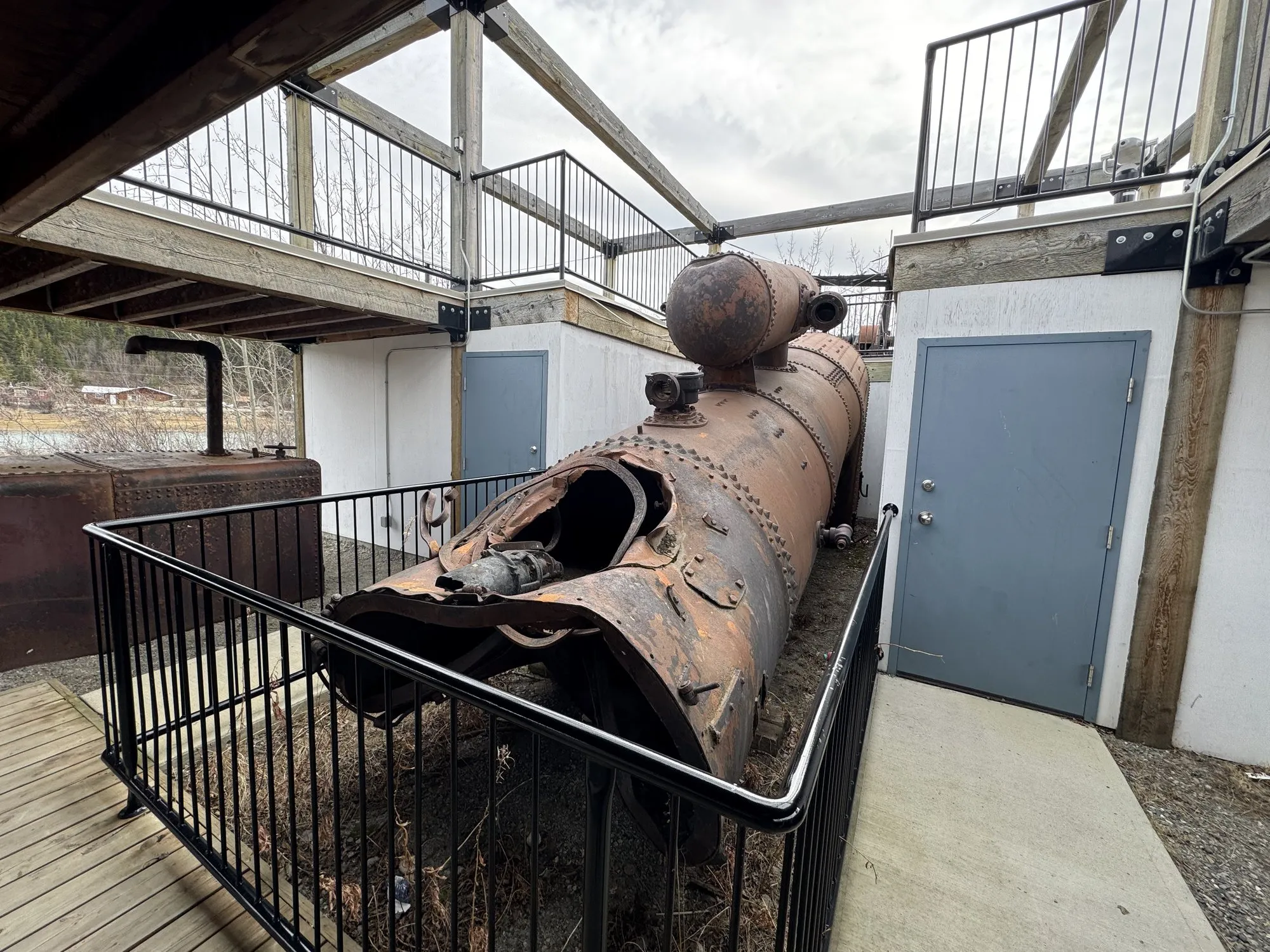 Rusted cylindrical boiler shell with its end opened, displayed beneath a deck