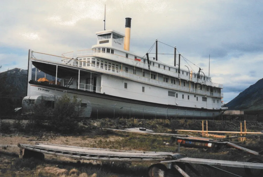 The Tutshi on blocking at Carcross in 1987, white hull and three decks with a buff and black stack, mountains behind